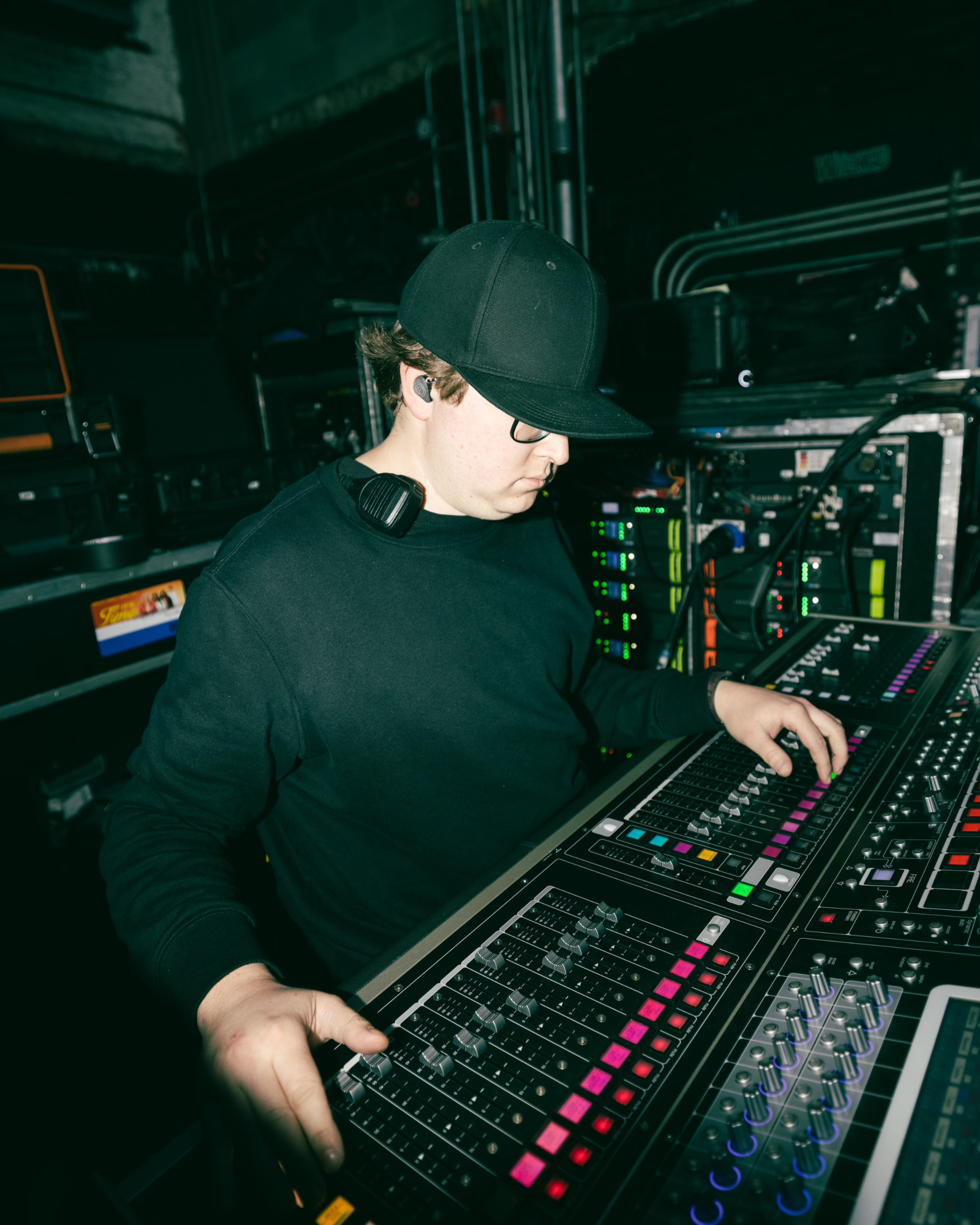 A man wearing IEMs and dressed in black adjusting controls on a mixing board.