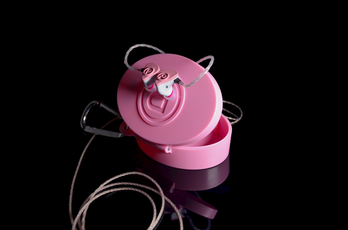 Pink oval IEM case with a hinged lid, shown open with a pair of wired in-ear monitors resting on top, photographed on a black reflective background.