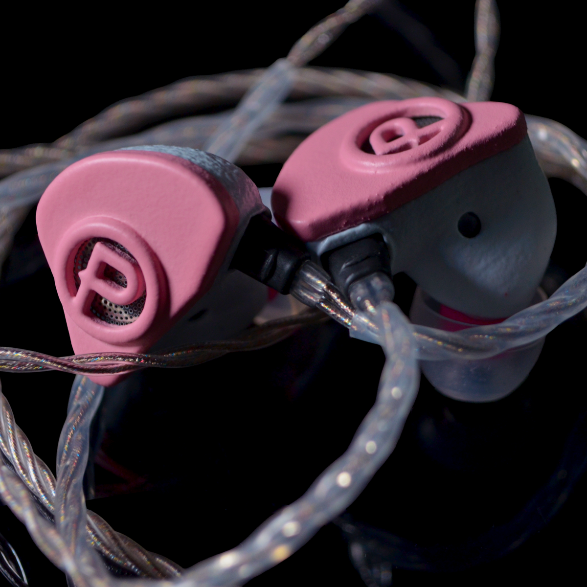 A close-up of two pink and white in-ear monitors tangled with clear braided cables against a black background. The earphones feature a circular 'P' logo on the faceplates.