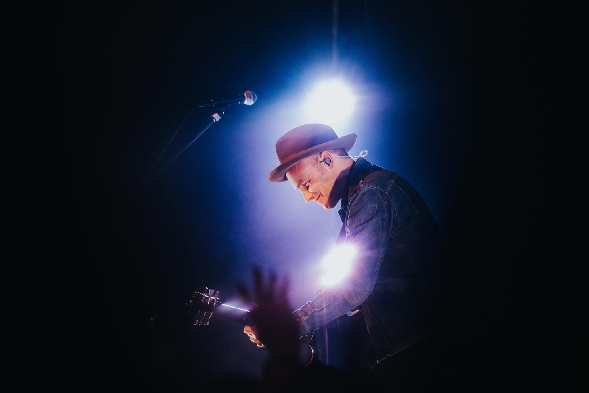 A musician wearing IEMs and a hat plays an acoustic guitar under a spotlight