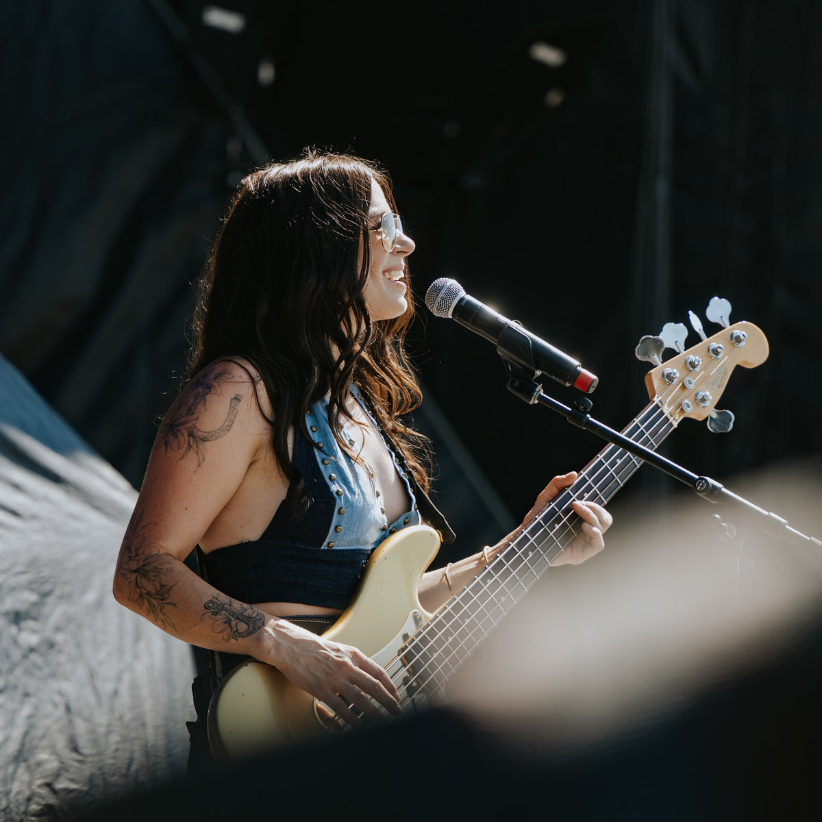A female musician performing live on stage, holding a light-colored bass guitar and singing into a microphone.