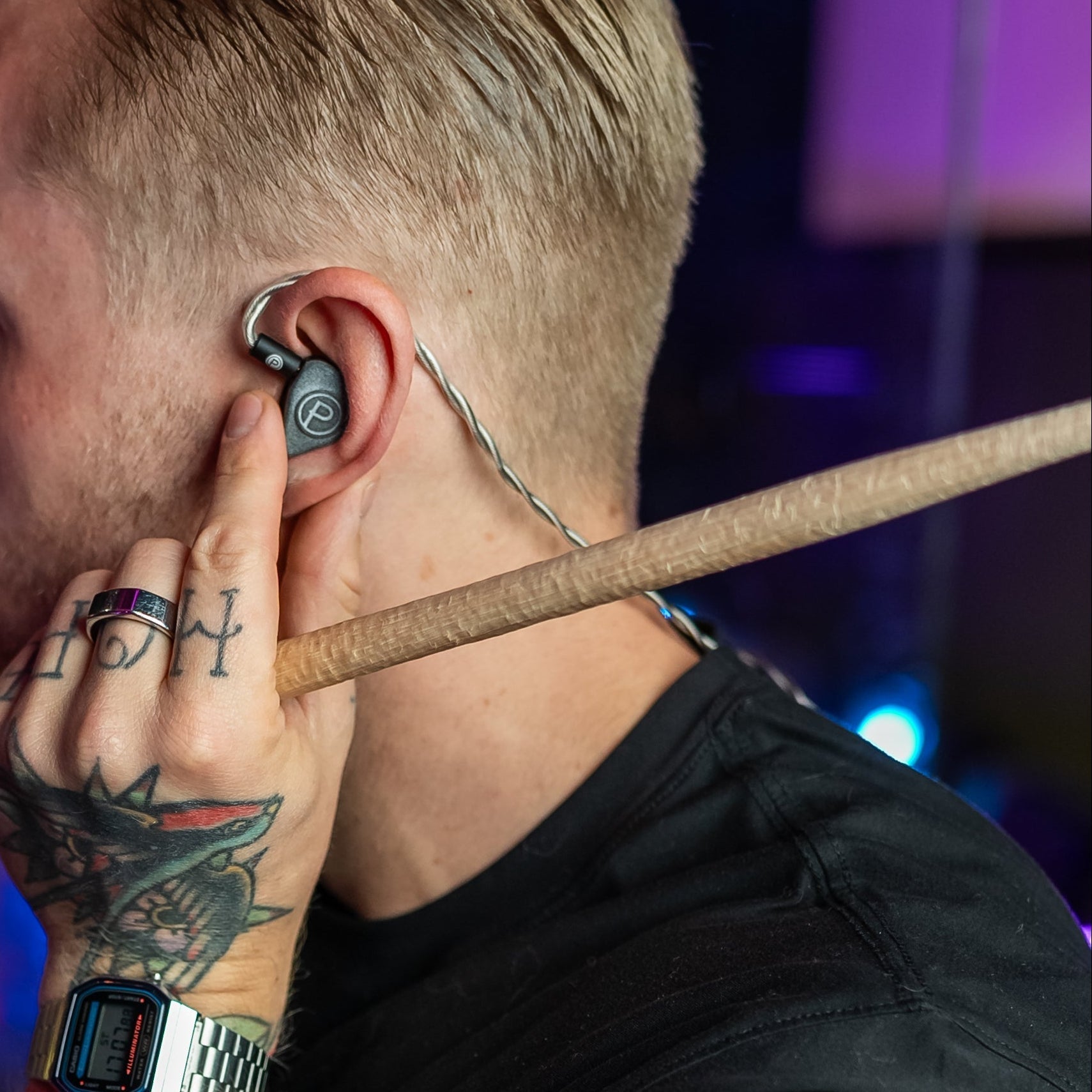 Person with tattoos holding drumsticks, wearing Unity IEMs in a dimly lit room.