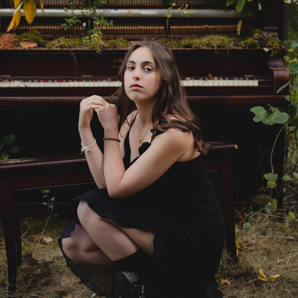 Kaiya Gamble in a black dress sitting in front of an old piano surrounded by greenery.