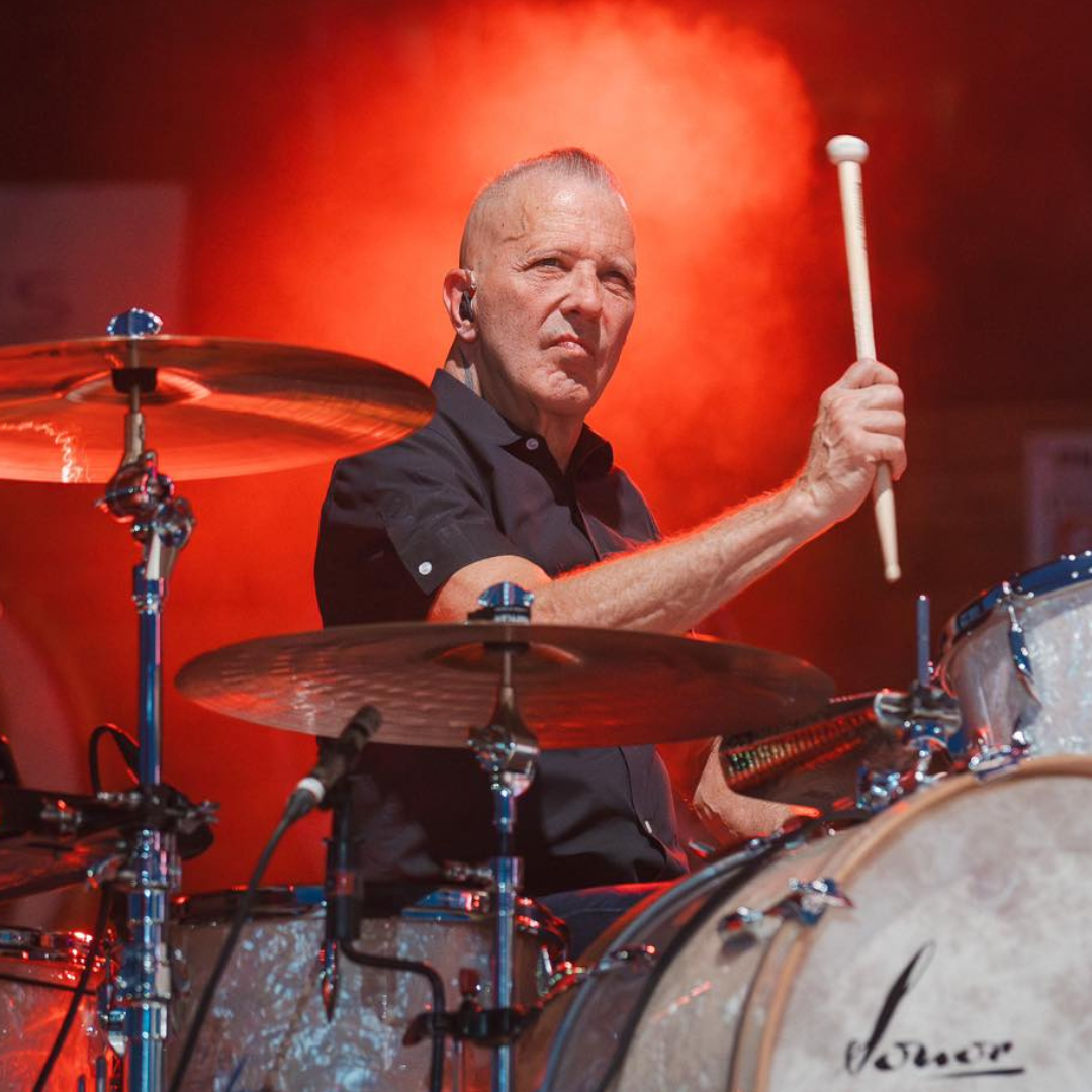 Paul Deakin of The Mavericks playing the drums with a red background.