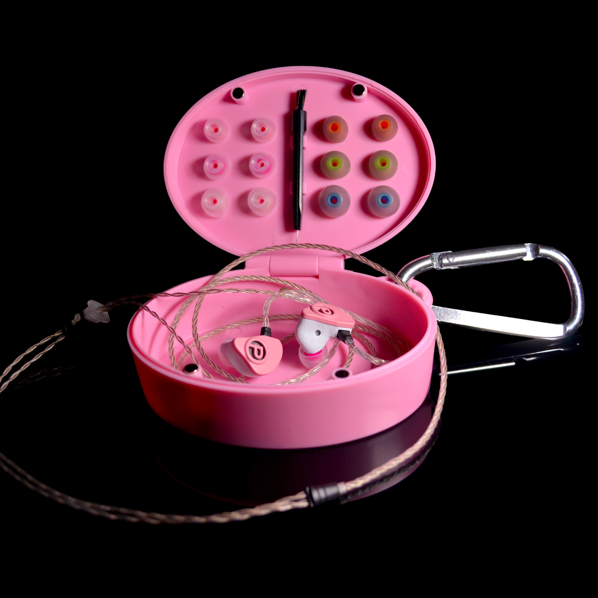 Pink in-ear monitors with a matching pink oval carrying case, shown open on a black background. The case contains multiple colourful silicone ear tips, a braided cable, and a silver carabiner is attached on the right.